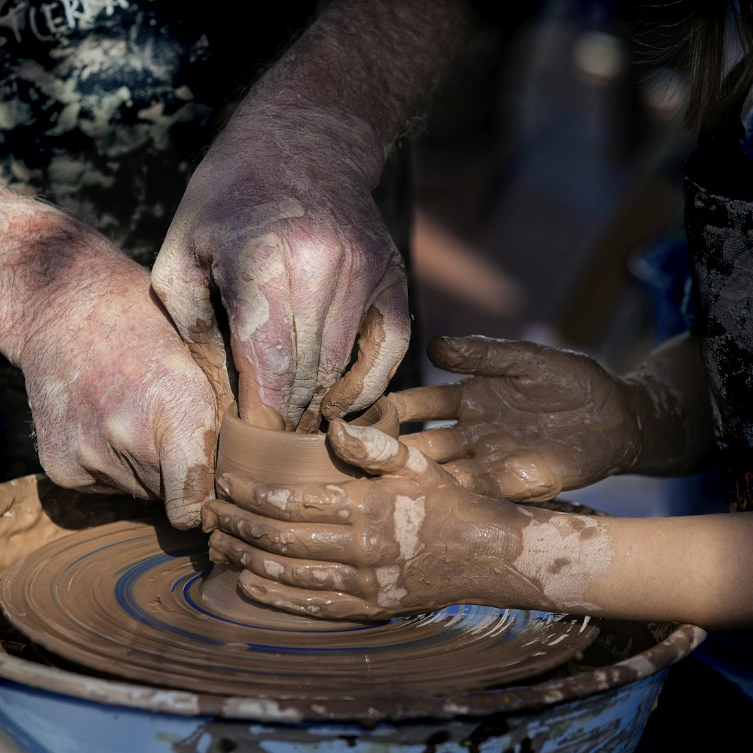 A master potter's hands guiding a child's hands as they shape one clay vessel together on the wheel